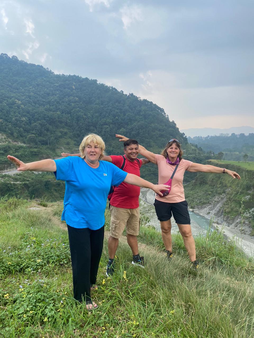 Three people posing playfully with arms outstretched on a grassy hill, scenic mountain background.